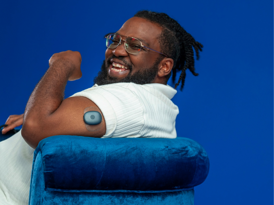 Eversense user smiling, flexing arm with a CGM sensor device attached, seated on a blue chair.